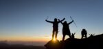 Silhouette of several hikers on the top of a mountain raising their arms in victory as the sun rises behind them.