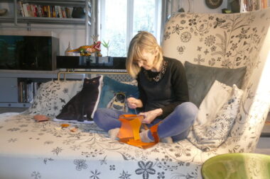 A woman sits cross-legged on an oversized chair, sewing with orange fabric.