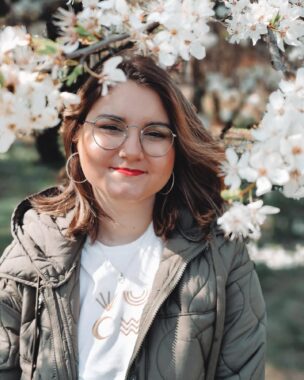 A woman with shoulder-length brown hair and glasses poses for a photo under a tree blooming with white flowers.