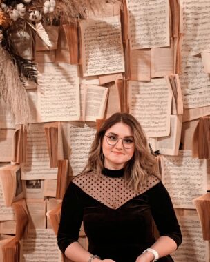 A woman in a black blouse smiles while standing in front of a wall, which appears to be covered in sheet music.