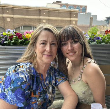A woman and her adult daughter sit outside in front of a raised bed of flowers and smile for a photo.