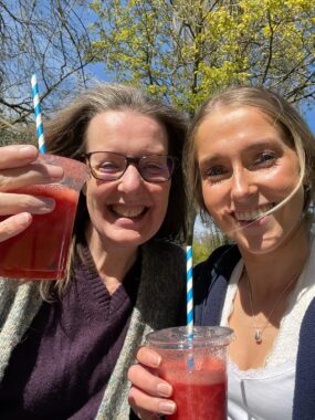 Two women, one older than the other, smile outside while holding up red drinks.