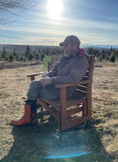 A man sits on a large wooden chair outside in the mountains. He's staring contemplatively off into the distance.