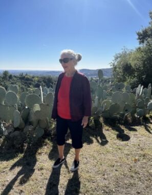 A woman in athletic clothing and sunglasses walks on a dirt path next to numerous cacti.