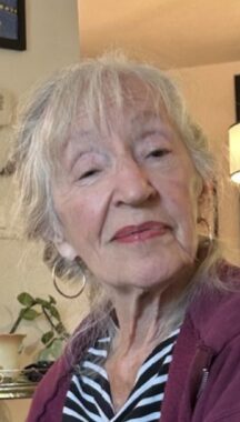 A close-up photo depicts an older woman tilting her head and smirking at the camera. She's wearing a striped blouse, a maroon jacket, gold hoop earrings, and lipstick. She appears to be sitting inside her home.
