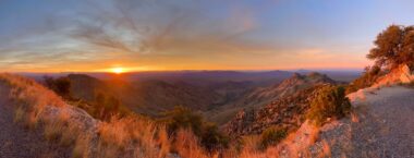 The sun sets just left of center in this picture, with orange sky lifting up into the blue. Beneath is what seems like desert terrain with some vegetation.