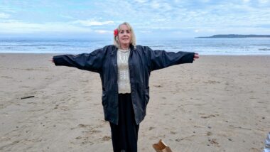 A woman with a red flower in her blond hair stands straight with her arms wide open to the side of her, as if in a Christ pose, while standing on a long beach that stretches to the back of the frame to the ocean. The blue of the ocean stretches out to the horizon and blends seamlessly with the blue of the sky, which is dotted with white, fluffy clouds. The woman is wearing a blue jacket.
