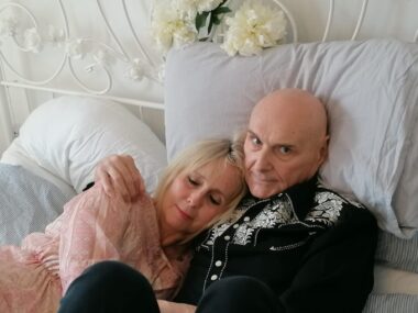A man and a woman embrace while lying down on a bed. White flowers are placed above the pillows. The man is wearing a Western-style button-up shirt and the woman a pink dress or blouse. The man looks directly into the camera while the woman has her eyes closed, indicating tenderness.