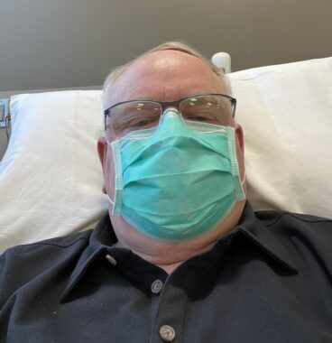 We see the head and shoulders of a man reclining on a hospital bed, though he appears in a dark gray-blue dress shirt. He wears glasses and a bright green mask covering his nose and mouth.