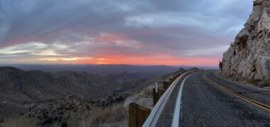 Photo by Sarah Jones A wide-angle photo shows a two-lane road curving around a mountain, with sloping hills in the distance. It's dusk, and the sunset has left a pink color near the horizon. The sparse vegetation gives the impression that the photo was taken somewhere in the desert, perhaps the American Southwest.