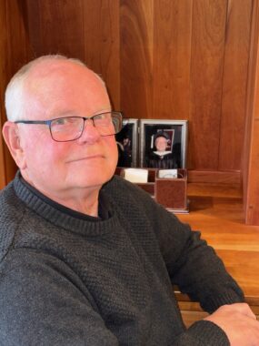 An older man in a dark gray sweater and glasses sits at a bright wood desk, on which we see pictures.