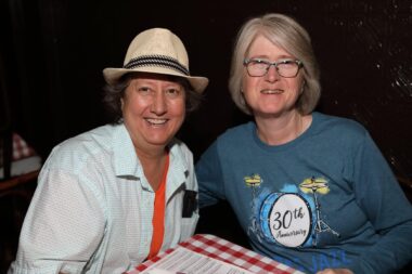 Two women sit next to each other at a restaurant dinner table. By the red-and-white checkered table covering, it's probably an Italian restaurant. The woman on the left is wearing a fedora.