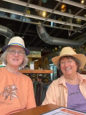 Two women sitting next to each other at a restaurant dinner table. They're both wearing hats and peach-colored shirts.