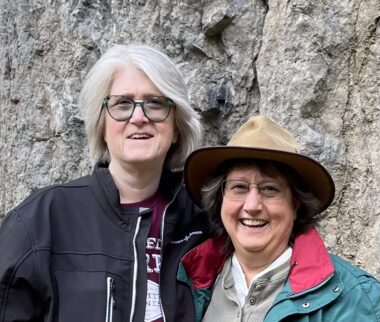 Two women stand in front of a large rock formation. They're both wearing thick-ish jackets, and one is wearing a hat.