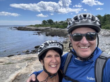 A man and a woman, clearly a couple, take a photo while hugging each other. They're both wearing bicycle helmets and are standing outdoors, in front of a body of water. The shore is lined with rocks and the sky is blue, with puffy, white clouds.