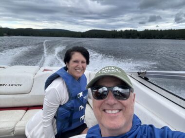 A man, who is taking a selfie, smiles giddily in the front of the frame, with a smiling woman behind him. They're both in a boat cruising along a river or lake. The man is driving and they're both wearing blue life jackets.