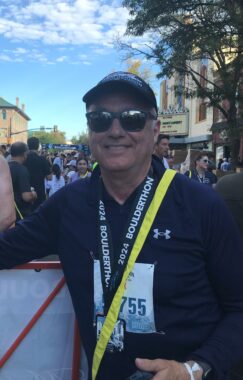 A small, vertical photo of a man leaning on something outdoors. He's clearly finished running a race and is wearing a baseball cap and sunglasses, as well as a bib number and a lanyard.