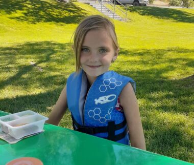 A young girl wearing a life jacket smiles while sitting at a picnic table in a large, grassy area with trees.