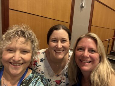 Three women smile for a close-up selfie in a large room with wood paneling on the walls.
