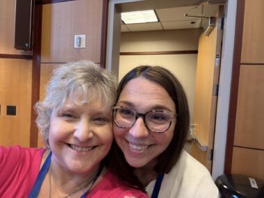 Two women smiles for a selfie in front of a doorway.